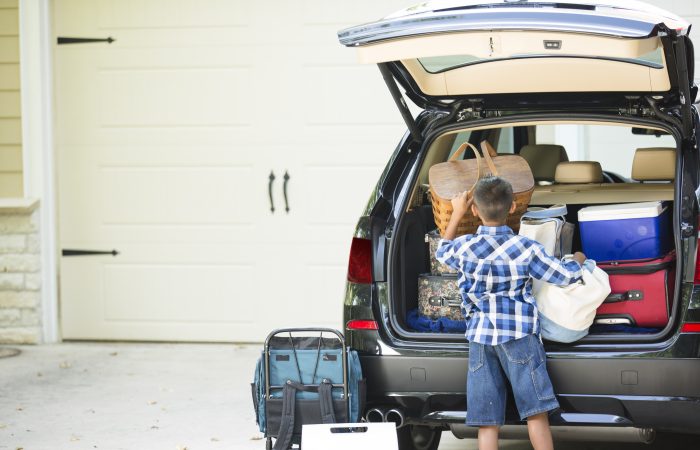 Family packs their vehicle for summer vacation.  One excited little boy waits by the car.